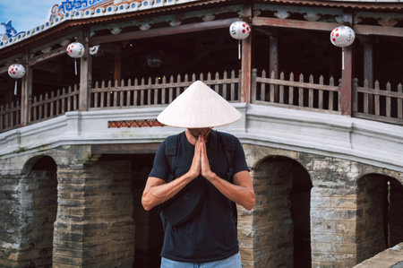 man traveler tourist in Vietnamese hat prays at the ancient Japanese covered bridge in old town in Hoi An in Vietnam in Asiaの写真素材