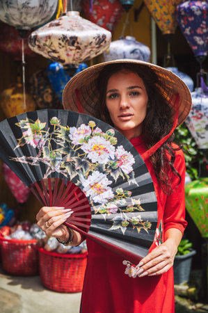 portrait of a beautiful tourist woman in Vietnamese cone hat Non La with fan in Hoi An in Vietnam in...
