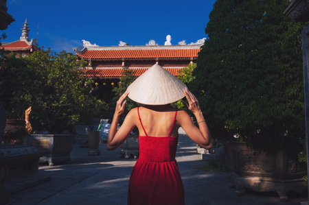 back of beautiful female traveler in a dress and a traditional Vietnamese hat Non La at a pagoda in...