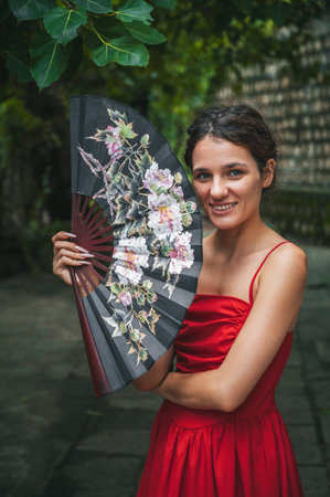 beautiful happy young woman in red dress with fan in the gardenの写真素材