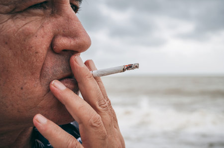 portrait of a sad adult elderly Caucasian man smoking a cigarette alone outdoors on beach close-upの写真素材