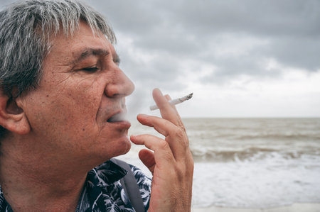 Lonely addicted sad elderly adult white man smokes a cigarette outdoors on the beach by seaの写真素材