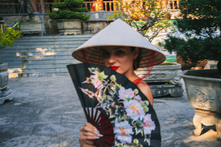 blurred portrait of a female tourist with a fan in a Vietnamese non la hat at a pagoda on trip to Vietnam in Asiaの写真素材