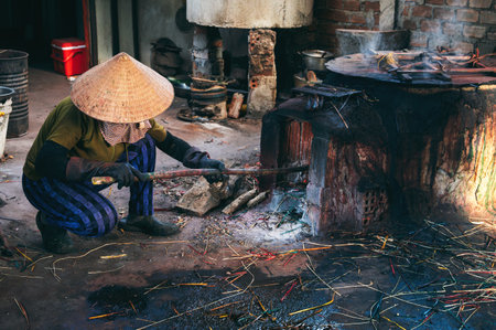 Vietnamese female stoker worker works in straw hat in a workshop and lights a coal furnace at a reed dyeing plant in Vietnam. Woman does dirty hard work in a factory in Asiaの写真素材