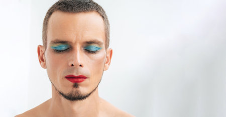 portrait of a beautiful feminine gay man with makeup and cosmetics on face. transgender male with red lipstick and eye shadow sparkle on eyes on white background with copy spaceの写真素材