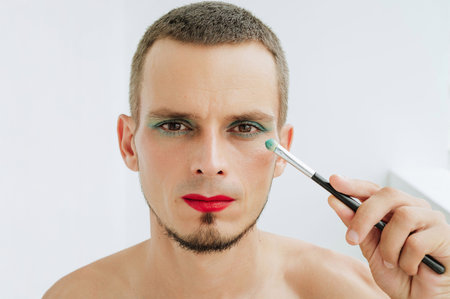 portrait of adult gay man with makeup and cosmetics on face. A handsome transgender male with red lipstick applied eye shadow sparkle on eyes with a brush on a white backgroundの写真素材