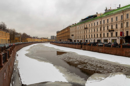 A quiet winter day in Saint Petersburg reveals a canal partially covered with ice and snow, flanked by historic buildings under an overcast sky.の写真素材