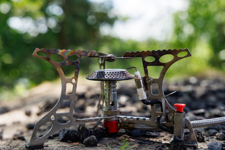 A small, compact camping stove sits on a bed of dirt and charcoal, with a lush green forest blurred...