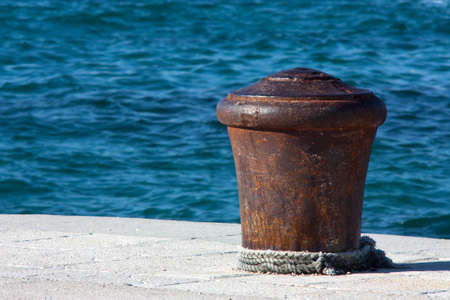 Big old rusty bollard on a stone pier, with a rope around it, and wavy blue sea in the backgroundの写真素材