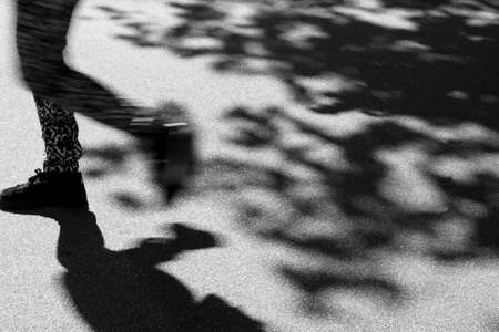 Legs of a walking person in motion blur and shadows of pedestrian and treetop foliage on city street pavementの写真素材