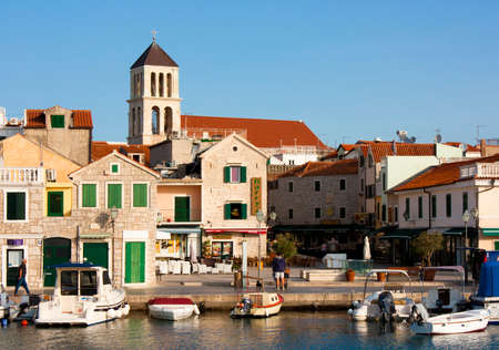 Vodice, Croatia - August 22, 2017: Sunny morning in small Mediterranean town square with stone houses and old church tower. with moored boats on the dock, and few people walking byのeditorial素材