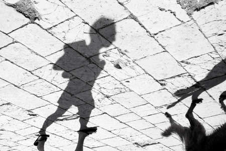 Blurry shadow of a person holding a book and walking a dog on  weathered ancient stone street promenade on Mediterranean island in summer sunの写真素材