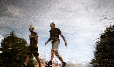Blurry reflection silhouette of two young men walking city streets , low angle view, in the puddle on a rainy summer dayの写真素材