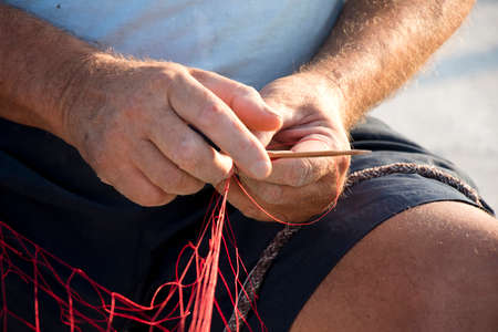 Fisherman hands repairing his red fishing net with needle , close up detail