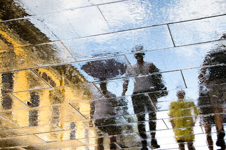 Blurry reflection shadow silhouette in a puddle of people walking wet sunny city street after rainの写真素材