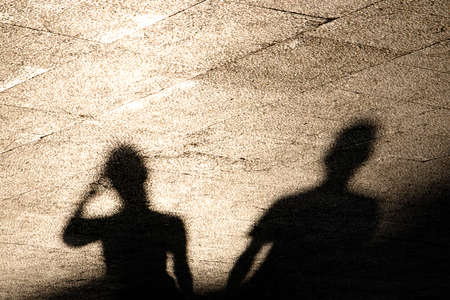 Blurry reflection shadow of two people on summer shiny sunny sidewalk in sepia black and whiteの写真素材