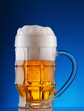 Faceted mug of light beer with foam on blue backgroundの写真素材