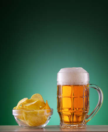 Faceted mug of light beer with foam and chips in a plate on a green backgroundの写真素材