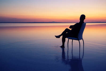 Silhouette of a man sitting on a chair in the middle of the lake at sunset. Calm water, dramatic...