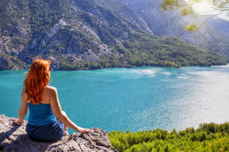 Girl with red hair on cliff top of canyon with green water. Summer landscape. Concept of solitude travel and harmonyの写真素材