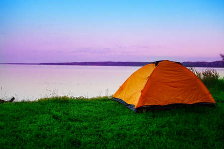 Tourist tent on the shore of a calm lake in the early morning. Pink dawn sky. Summer landscape. The concept of travel, privacy, freedom.の写真素材
