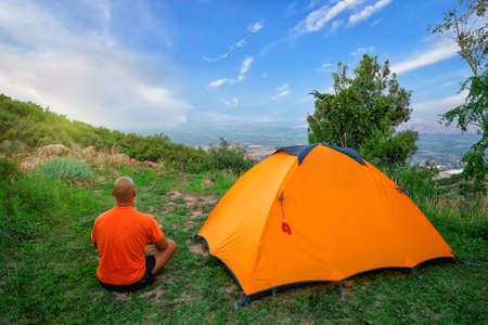 Man in an orange T-shirt sits near a tent on a hill. Blue sky, bright sun.の写真素材