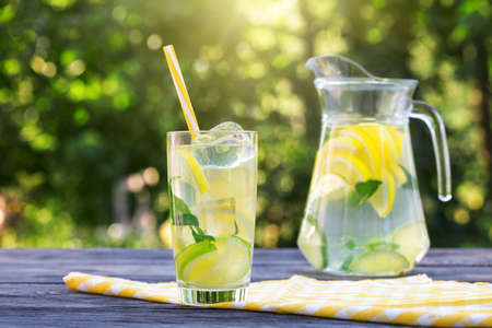 Lemonade with ice in glass with straw and jug on wooden tableの写真素材