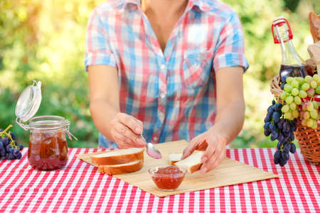 A woman in a plaid shirt spreads jam on bread. Red tablecloth, picnic basket, grapes. Sunny day. Picnic conceptの写真素材