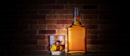 Glass of whiskey with ice and bottle on a dark brick background