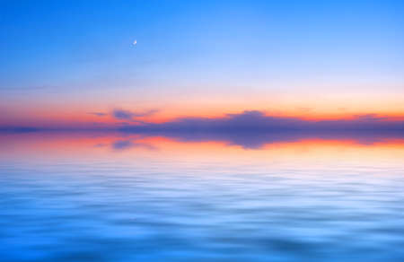 Sunset sky panorama with young moon above water surfaceの写真素材