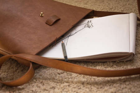 From the leather brown bag you can see an album for drawing with a non-drawn picture and a pencil. The bag is prepared for the trip and the most important things are collected for the master.の写真素材