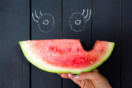 A piece of a bitten and juicy watermelon on a background of a wooden surface of black color. On the surface are painted eyes, in combination with a watermelon you get a funny face.の写真素材