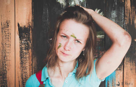 Beautiful and young girl standing on the background of painted wall paint. The girl's face was stained with a green paint - she was painting with a brush. With her left hand, she smoothes her hair back.の写真素材