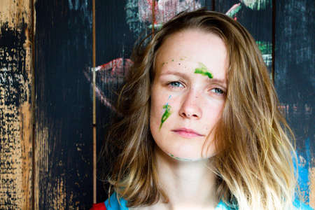 Beautiful and young girl standing on a wooden wall background. The girl's face is stained with green paint.の写真素材