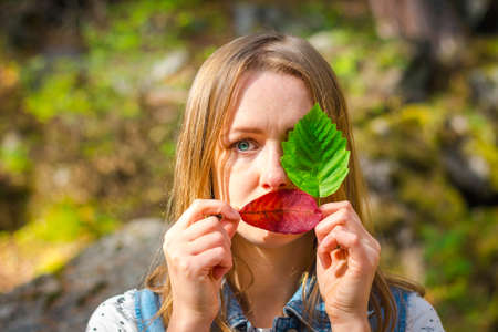Beautiful and young girl holds leaves. With leaves, she covered one eye and her delicate lips. A girl stands on a background of nature.の写真素材