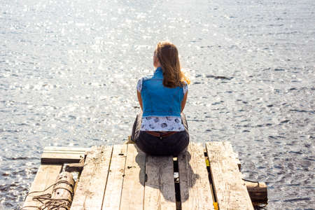 A beautiful and young girl is sitting on a pier. She looks at the clear water of the lake and dreams. The sun's rays illuminate the water and everything around.の写真素材
