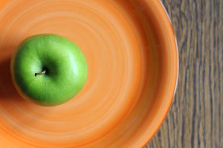 Green apple on an orange plate. The plate is made of clay and stands on a wooden surface. The apple is fresh and ready to eat.の写真素材