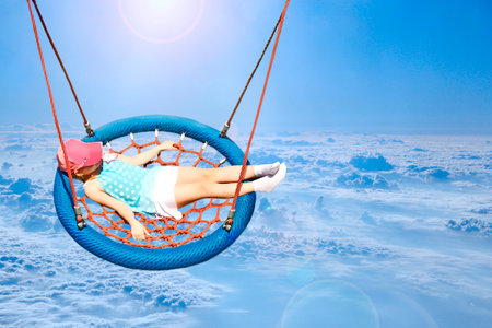 Flight over clouds. Little girl playing on swing. Flying over white clouds. Beautiful panorama with heaven. little girl riding in hammock above clouds. Girl soars above the sky. white cloud belowの写真素材