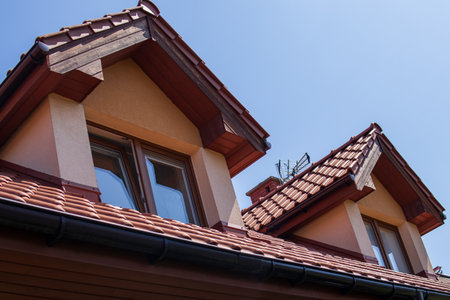 Close-up view of a house roof with dormer windows and red ceramic tiles, set against a clear blue...
