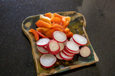 Sliced fresh radishes and orange bell pepper on a rustic handmade ceramic plate, placed on a black countertop. A simple, healthy vegan snack idea.の写真素材