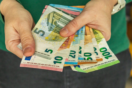 Close-up of hands holding multiple euro banknotes in mixed denominations, illustrating cash flow, financial operations and monetary handling.の写真素材