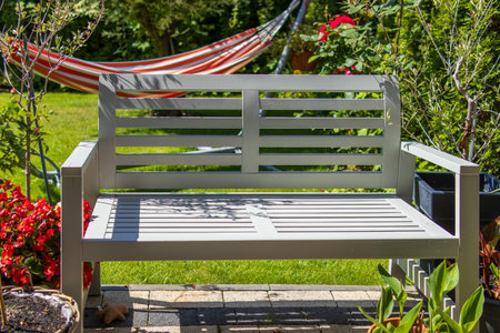 A sunlit garden bench surrounded by blooming flowers and greenery. A striped hammock swings gently in the background, evoking a calm, relaxing atmosphere in a cozy backyard.の写真素材