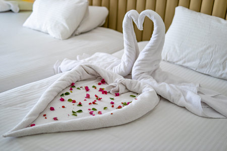 Romantic towel swans forming a heart shape on luxury hotel bed with flower petals. Honeymoon, wedding travel, love and hospitality concept with cozy bedroom interior.の写真素材
