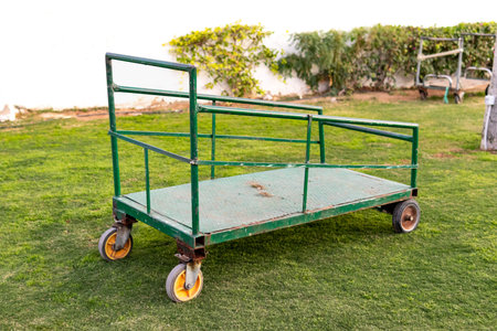 Empty green metal utility cart with wheels standing on grass outdoors. Industrial transport trolley...