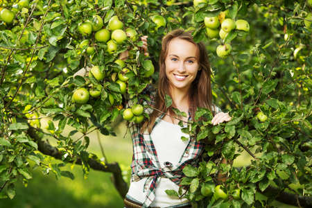 Woman in an apple tree garden during the harvest season. Young smiling beautiful woman is standing among the apple trees with ripe organic apples on it. Healthy country lifestyle concept.の写真素材