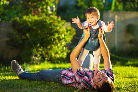 Father and baby daughter are playing outdoors. Young daddy and his cute little baby-girl are having fun in the sunny garden. Happy childhood and parenthood concept. Focus on the daughter.の写真素材