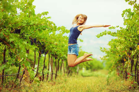 Happy woman in a vineyard is jumping and having fun during the harvest season. Fresh ripe grapes on a vine at the background. Eco lifestyle concept.の写真素材
