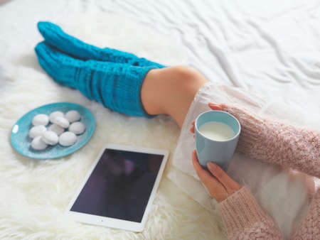 Woman using tablet at cozy home atmosphere on the bed. Young woman with cup of milk in hands...