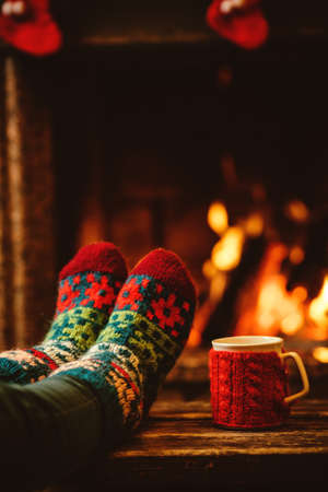 Feet in woollen socks by the Christmas fireplace. Woman relaxes by warm fire with a cup of hot drink...