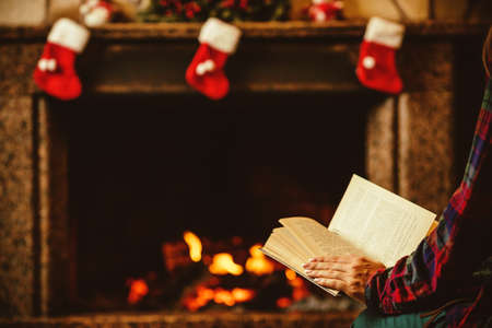 Woman reading a book by the fireplace. Young woman reading a book by the warm fireplace decorated for Christmas. Relaxed holiday evening concept.の写真素材
